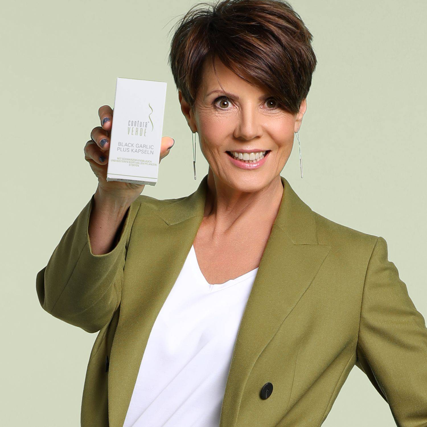 Eine Frau hält eine Verpackung mit der Aufschrift 'BLACK GARLIC PLUS KAPSELN' in der Hand. Sie trägt einen olivgrünen Blazer und eine weiße Bluse.