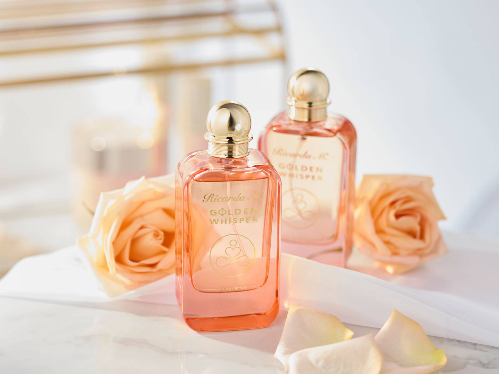 Zwei Flaschen Ricarda M. Golden Whisper Parfüm in rosa Glas mit goldfarbenem Verschluss, umgeben von hellrosa Rosenblüten auf weißer Oberfläche.