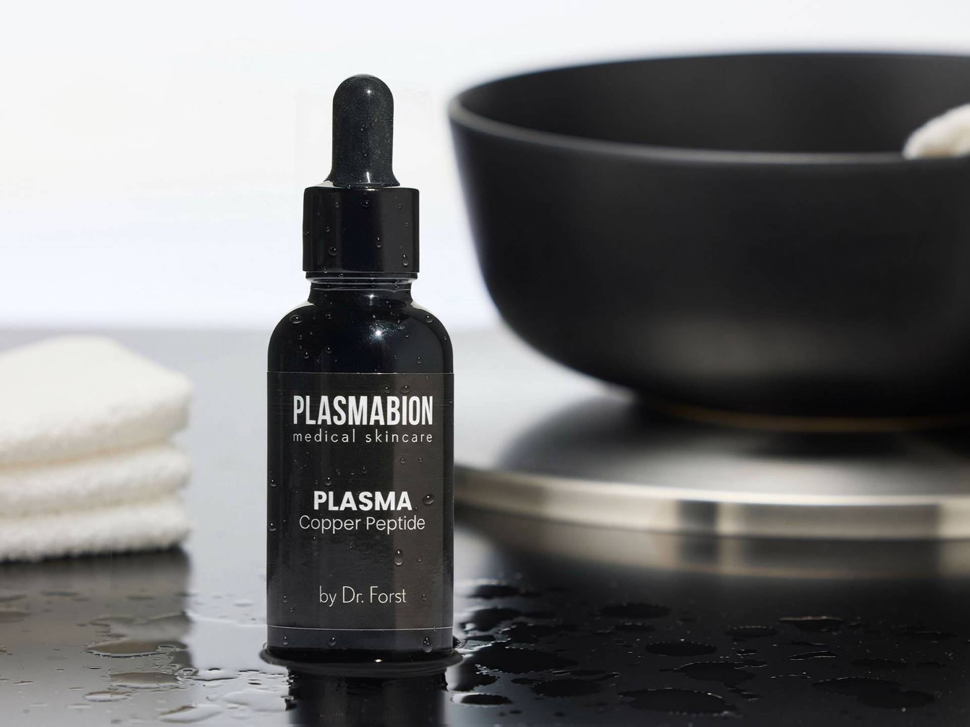 Schwarze Flasche mit Pipette, beschriftet mit 'PLASMABION medical skincare PLASMA Copper Peptide by Dr. Forst', steht auf nasser Oberfläche neben weißen Handtüchern und schwarzer Schale.