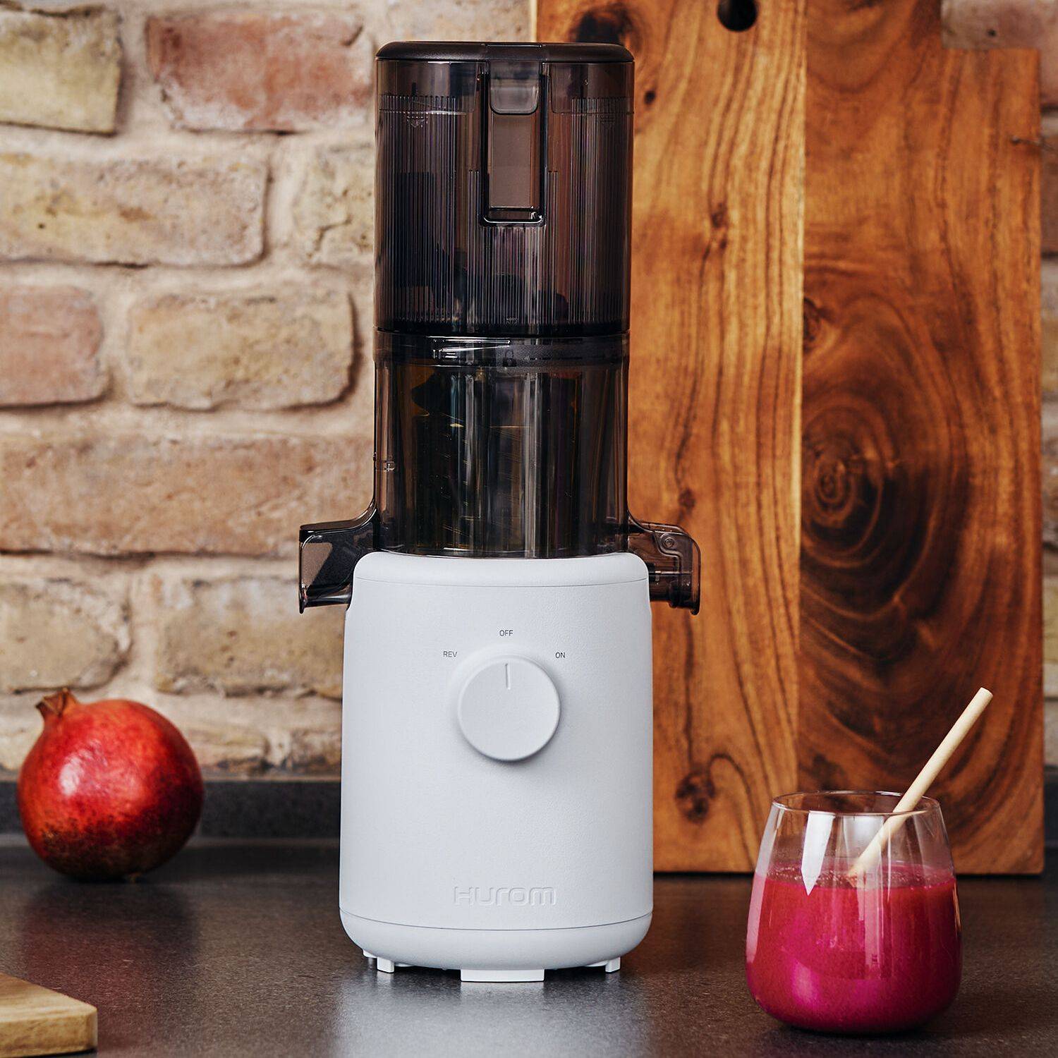 Weißer Hurom-Entsafter mit transparentem Behälter, daneben Granatapfel und Glas mit rotem Saft und Holzrührstab auf Küchenarbeitsplatte.