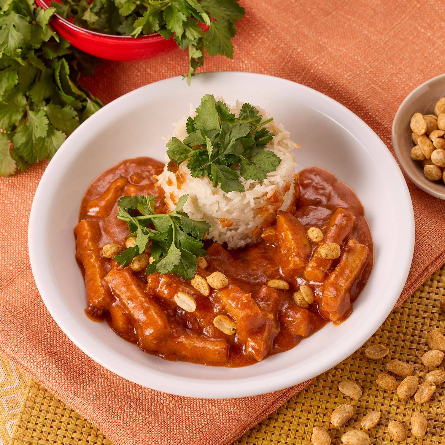 Weißer Teller mit Hähnchencurry in roter Sauce, garniert mit Erdnüssen und Koriander, dazu eine Portion Reis mit Korianderzweig.