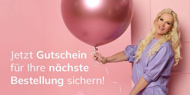 Frau mit langem, gewelltem Haar hält großen, runden Luftballon vor rosa Wand, trägt lilafarbenes Kleid mit Puffärmeln 