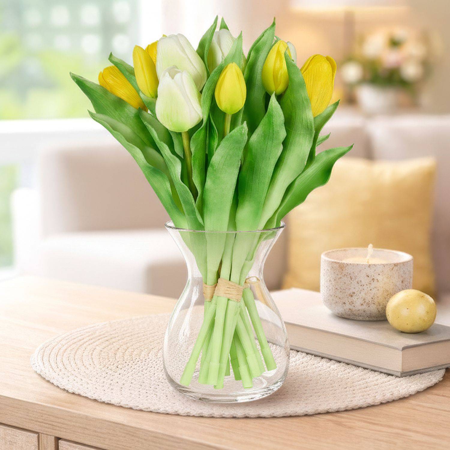 Strauß Tulpen in einer Glasvase auf einem Tisch mit Buch, Kerze und Kugel vor einem Sofa mit Kissen und Decke im Hintergrund