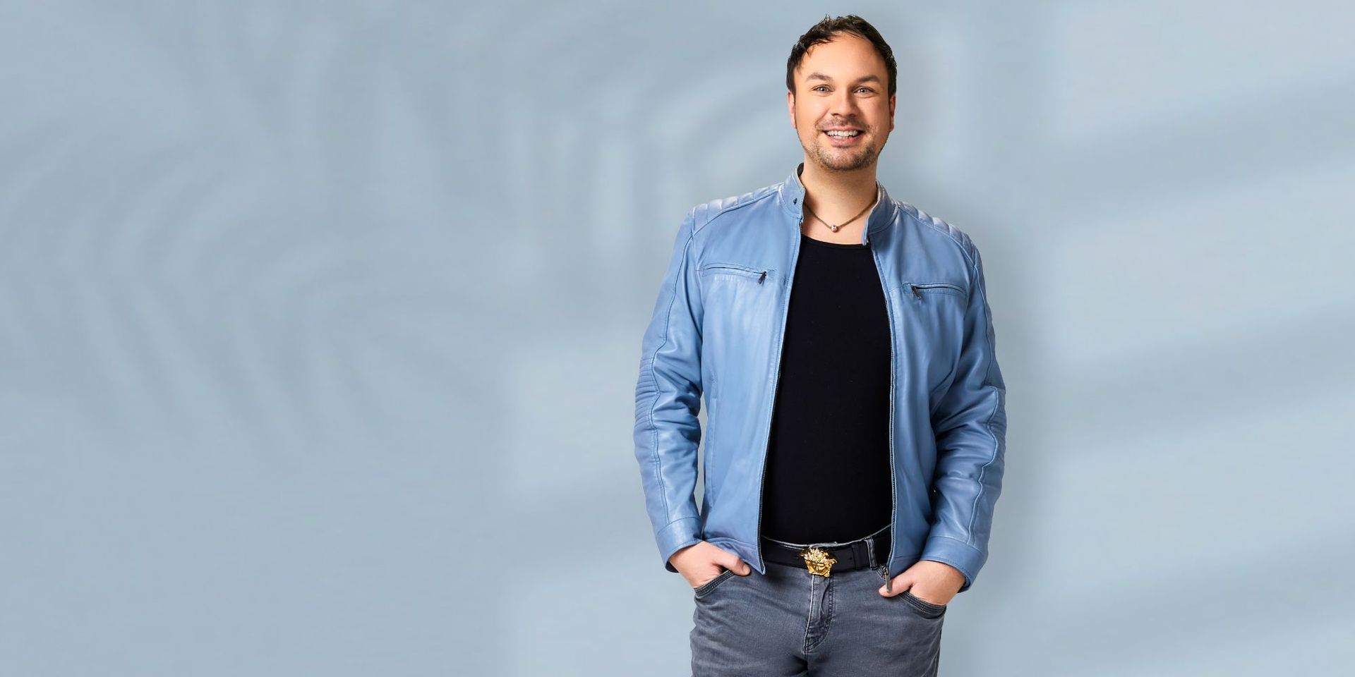 CHANNEL21 Experte Christian Materne lächelnd mit den Händen in den Hosentaschen stehend vor einer blau-grauen Wand.