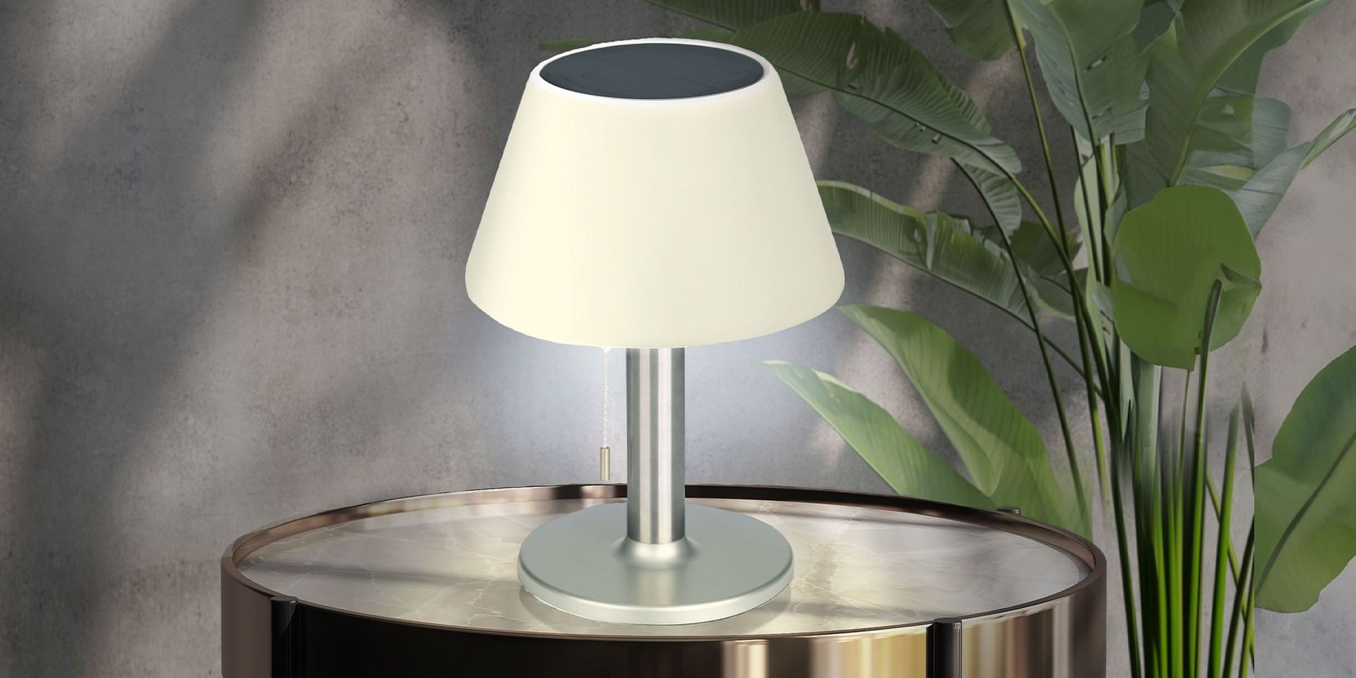 Solarbetriebene Tischlampe mit weißem Schirm auf einem runden Tisch vor grünen Blättern.
