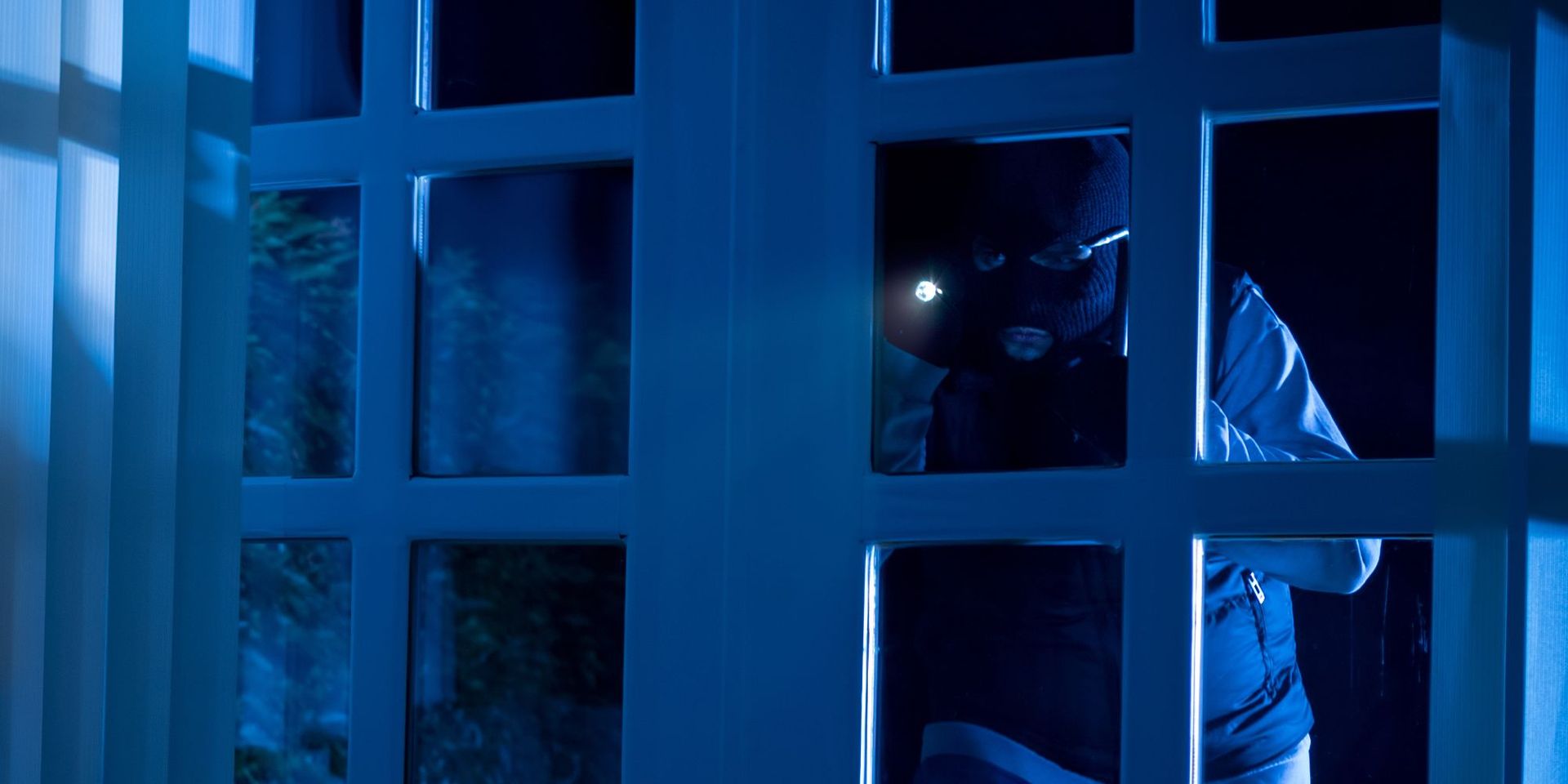 Einbrecher steht vor einem Fenster und leuchtet mit einer Taschenlampe herein
