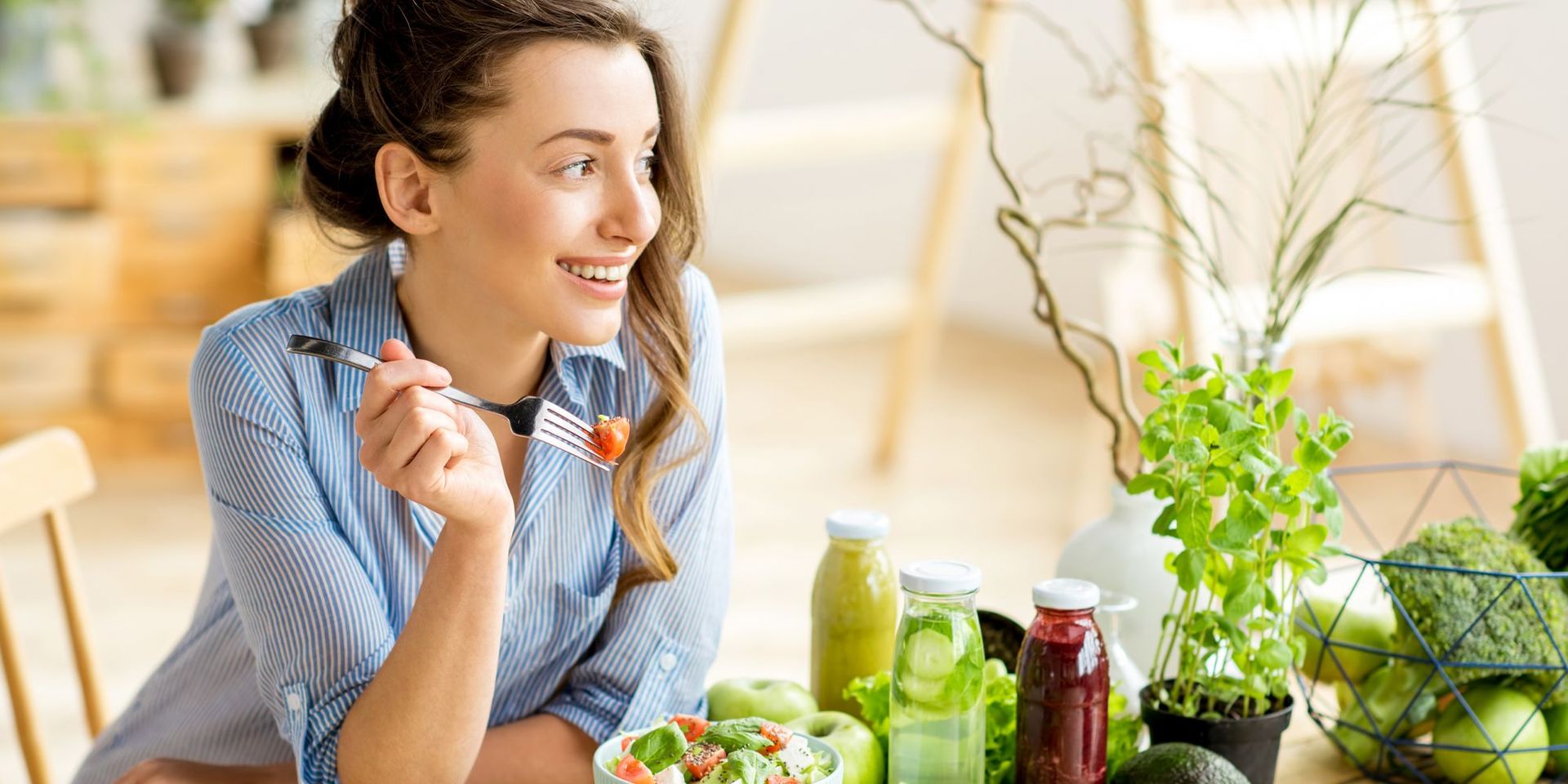 Junge Frau sitzt am Tisch, welcher mit Gemüse und Smoothies gedeckt ist, und isst eine Erdbeere.