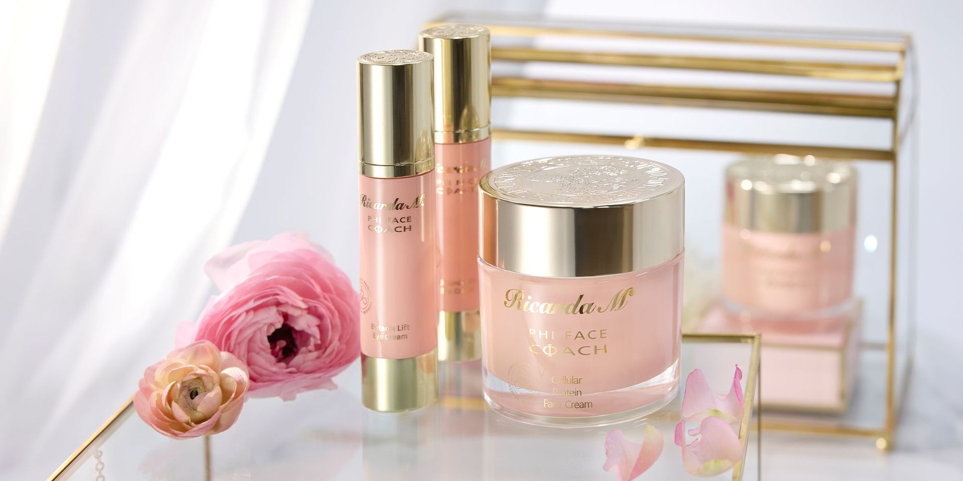 Drei rosa und goldene Kosmetikprodukte der Marke 'Ricarda M.' auf einem Spiegel mit rosa Blumen dekoriert