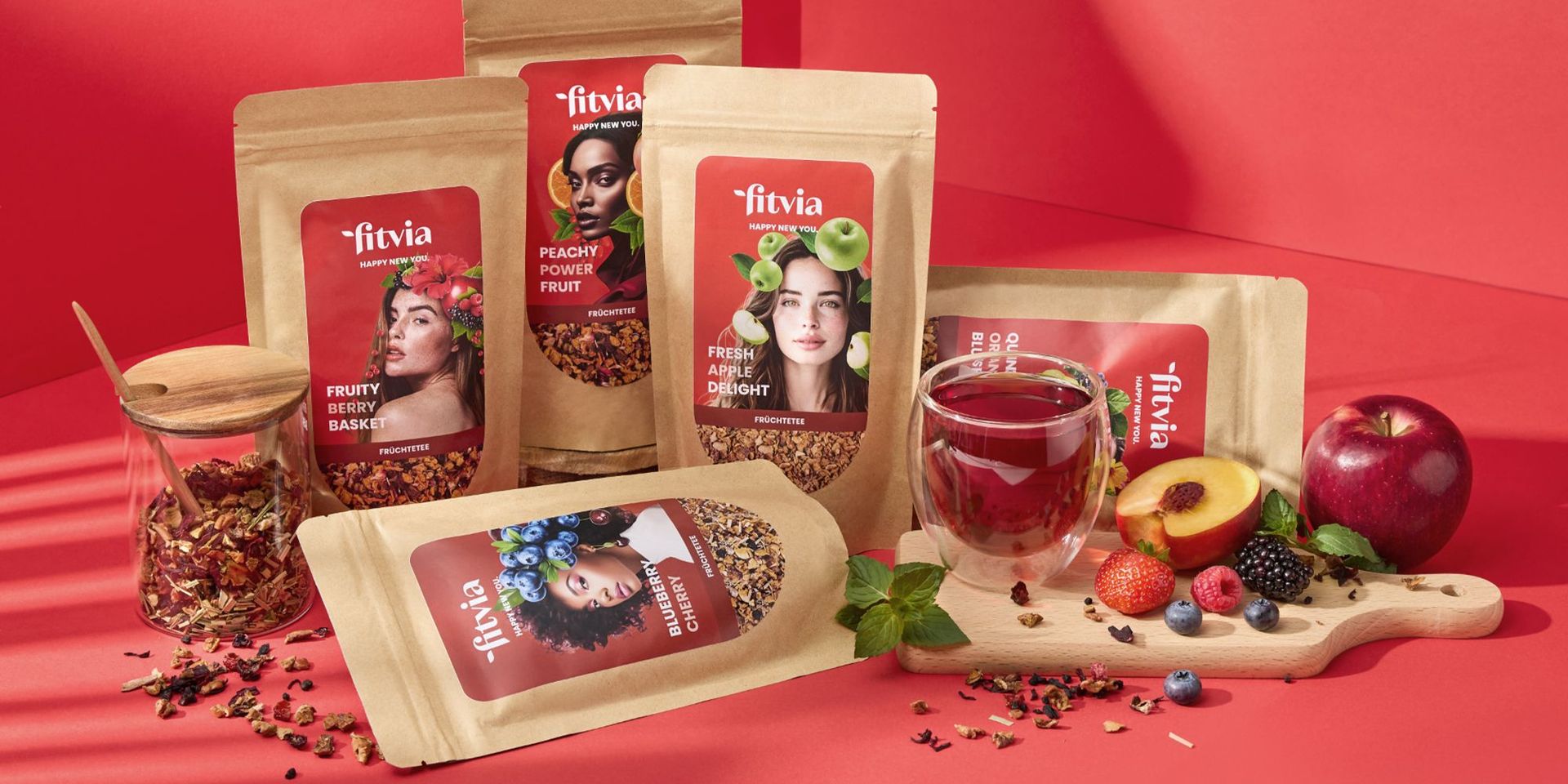 Fitvia Früchtetee-Packungen auf rotem Hintergrund mit frischen Früchten und Teeglas