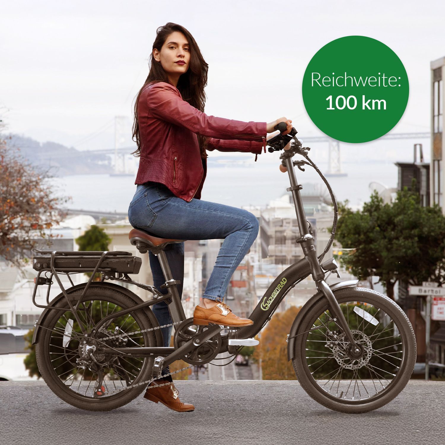 Frau in roter Jacke und blauen Jeans sitzt auf einem schwarzen Elektro-Klapprad mit braunem Sattel, im Hintergrund Stadt und Brücke, daneben gefaltetes Klapprad