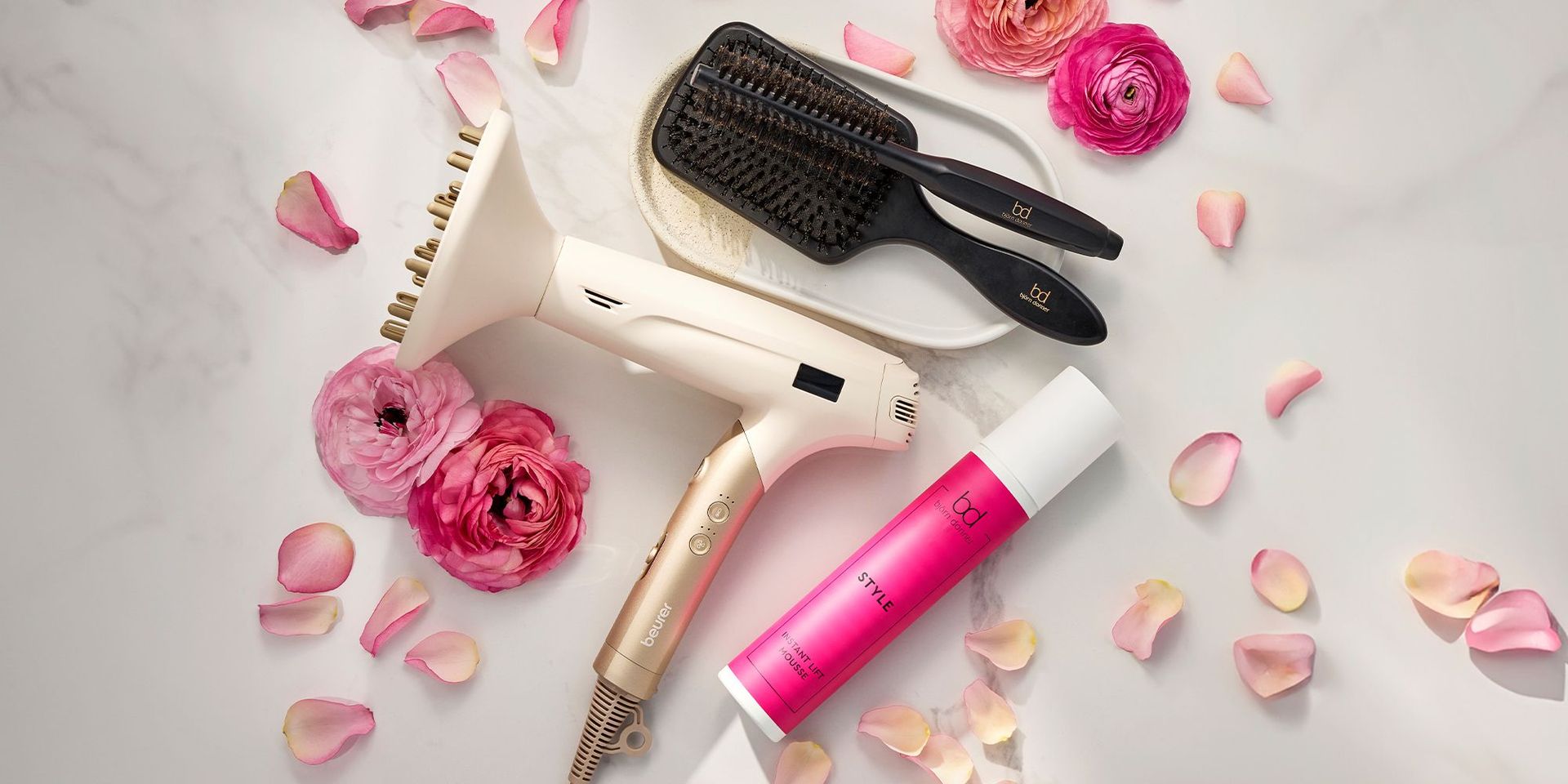 Ein Haartrockner, ein Haarstylingprodukt und eine Haarbürste liegen umgeben von Rosablüten auf einem hellen Hintergrund
