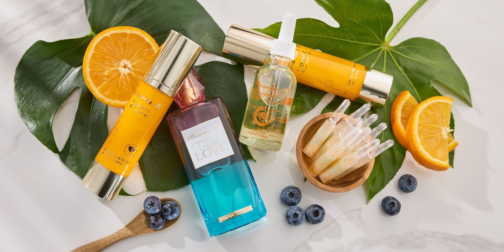 Zwei gelbe Pflegecremes, ein blauer Duftspray, ein Serum mit Pipette, Orangenscheiben, Heidelbeeren und grüne Blätter auf weißem Untergrund