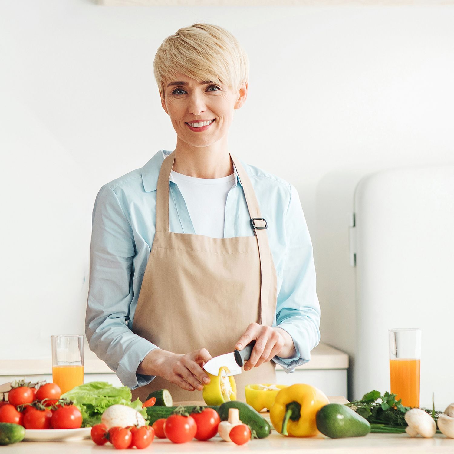Person mit hellblondem Haar trägt beige Schürze und schneidet gelbe Paprika in moderner Küche mit frischem Gemüse und Laptop