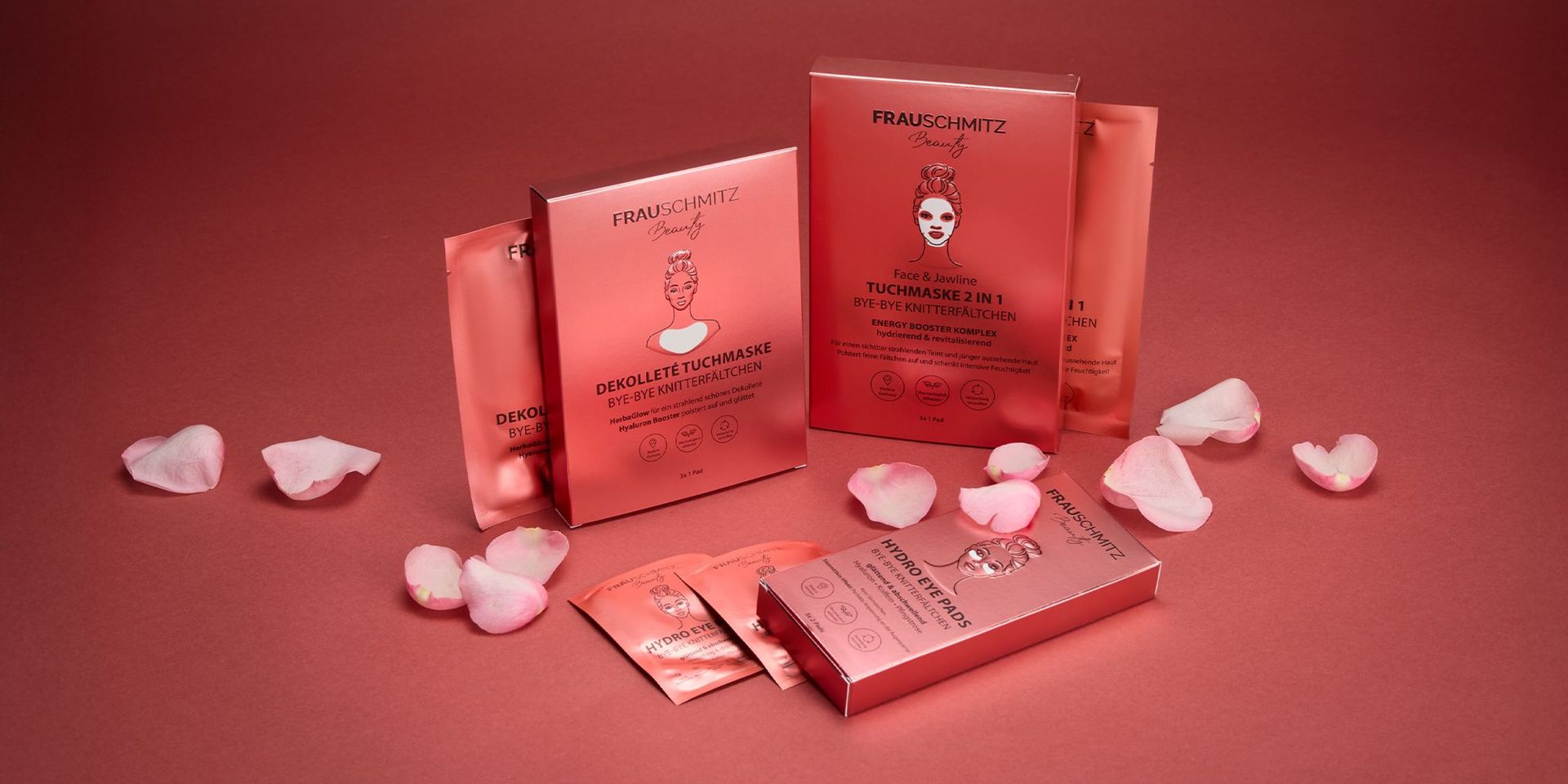 Mehrere rosa Verpackungen von FRAUSCHMITZ Beauty Produkten, darunter Dekolleté Tuchmaske und Face & Jawline Tuchmaske 2 in 1, umgeben von rosa Blütenblättern.