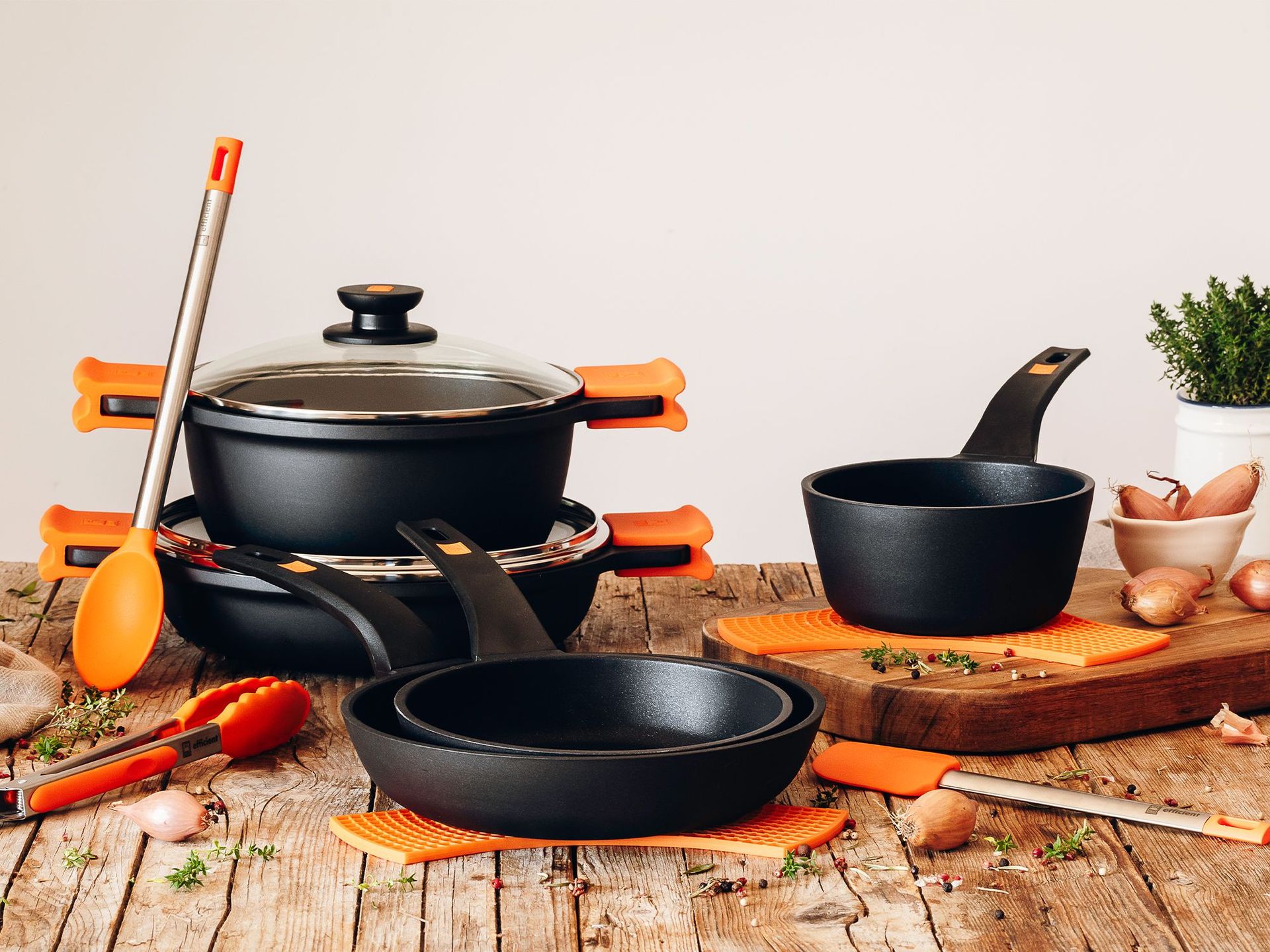 Schwarze Töpfe und Pfannen mit orangefarbenen Griffen auf Holzoberfläche, daneben Küchenutensilien mit orangefarbenen Griffen, Schalotten und ein Topf mit grünem Kraut.