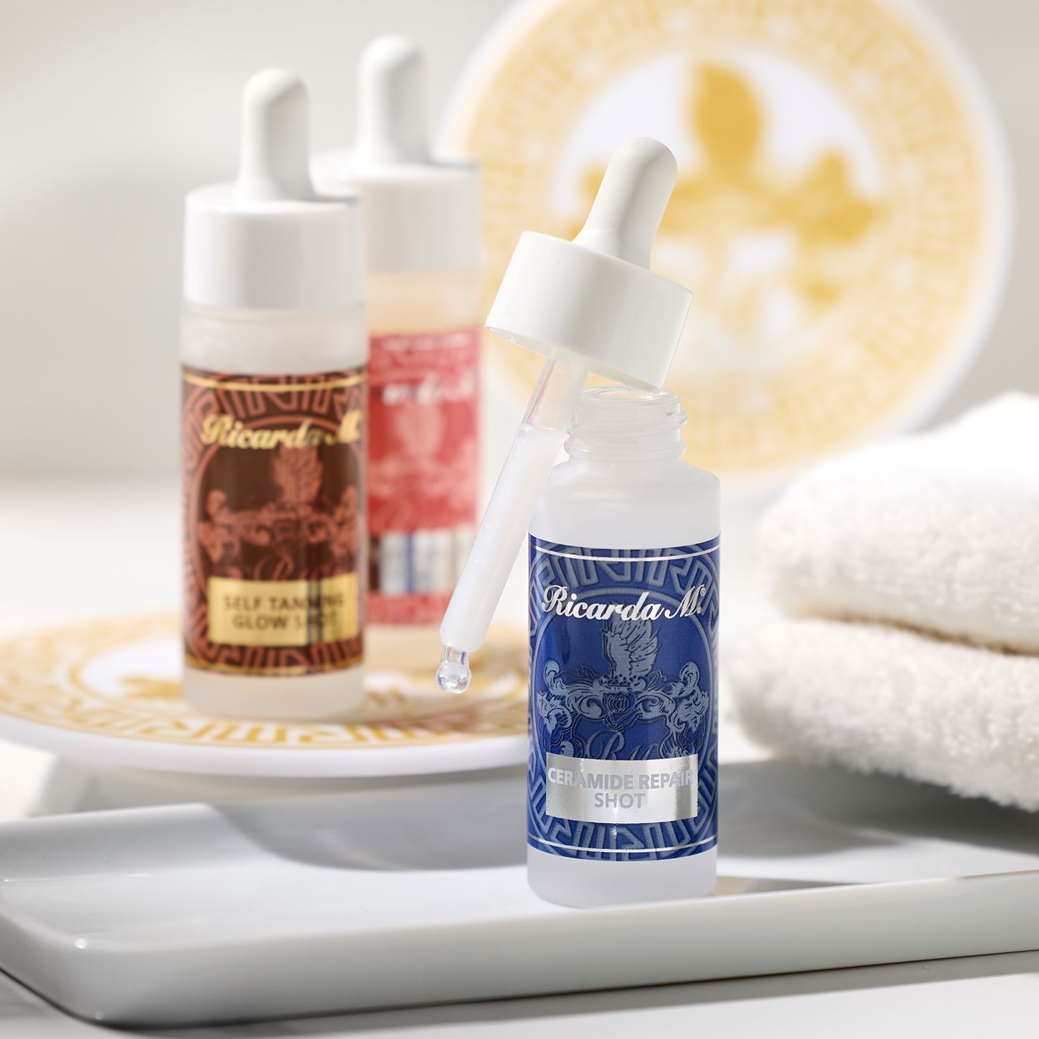 Drei Ricarda M Hautpflege-Shots in Flaschen mit Pipetten, vorn Ceramide Repair Shot in blauer Flasche, dahinter Self Tanner Glow Shot und ein weiterer Shot, daneben weiße Handtücher und gold-weiße Untersetzer.