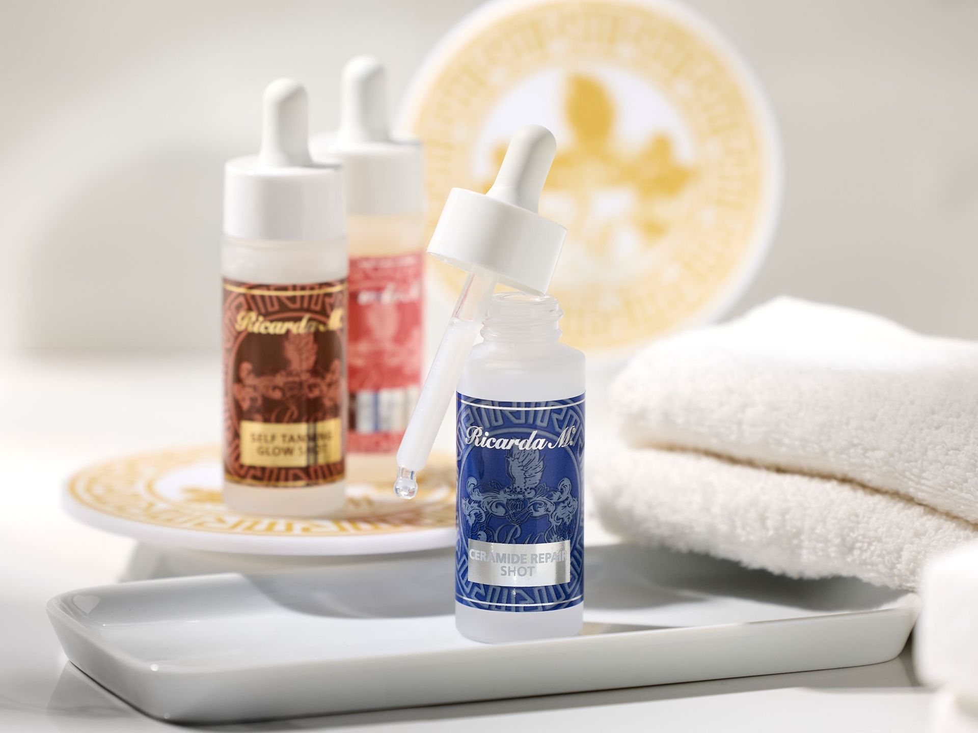 Drei Ricarda M Hautpflege-Shots in Flaschen mit Pipetten, vorn Ceramide Repair Shot in blauer Flasche, dahinter Self Tanner Glow Shot und ein weiterer Shot, daneben weiße Handtücher und gold-weiße Untersetzer.