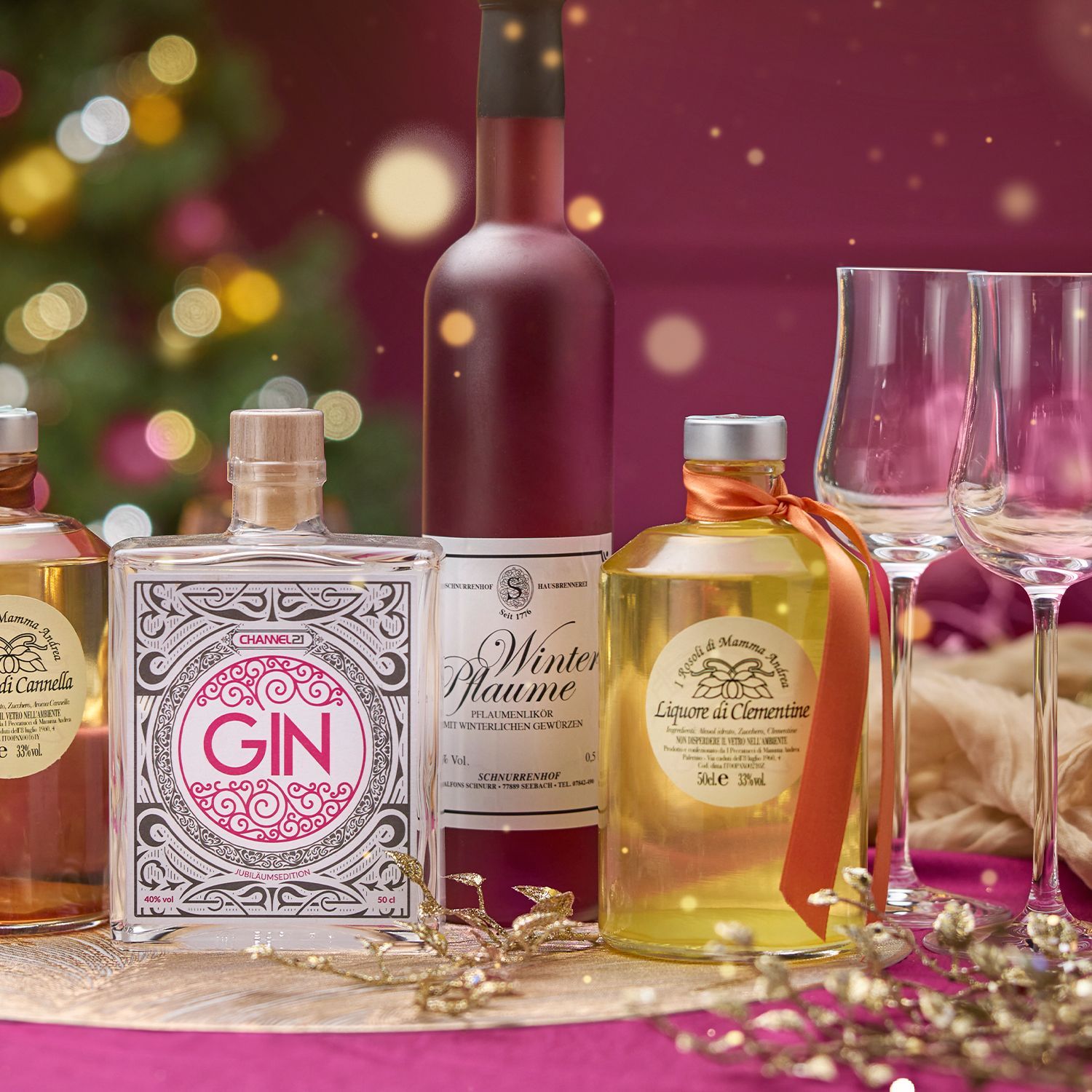 Verschiedene Flaschen mit Spirituosen und zwei leere Weingläser vor weihnachtlicher Dekoration mit Geschenk und Lichterkette.