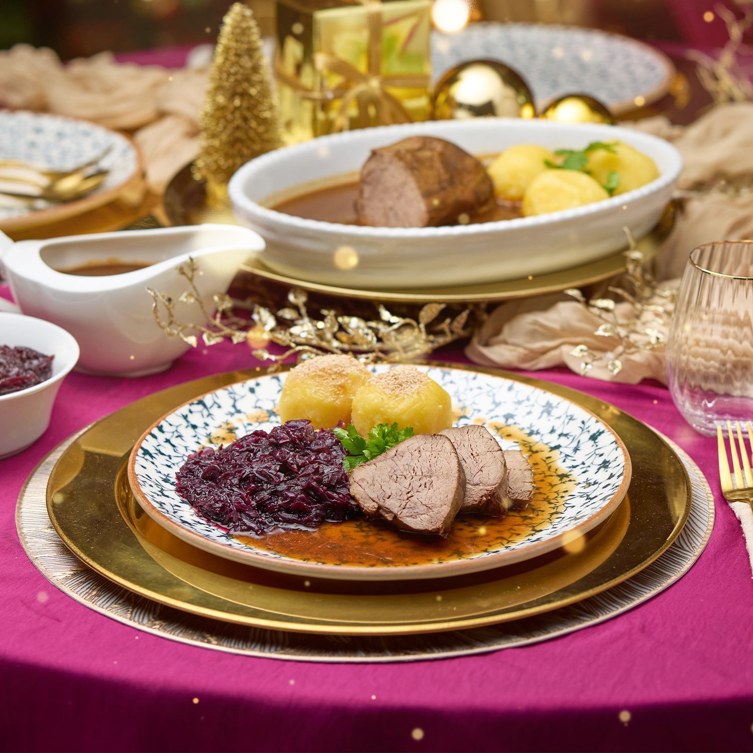 Festlich gedeckter Tisch mit goldfarbenem Besteck, Glasgeschirr, Servietten und Tellern mit Braten, Klößen und Rotkohl, umgeben von Weihnachtsdeko.