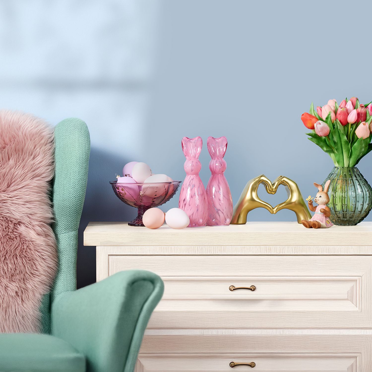 Heller Nachttisch mit rosa Plüschkissen auf grünem Sessel, darauf rosa Ostereier in Glasschale, zwei rosa Hasenfiguren, goldene Hände, die ein Herz formen, kleine Hasenfigur und Vase mit Tulpen.