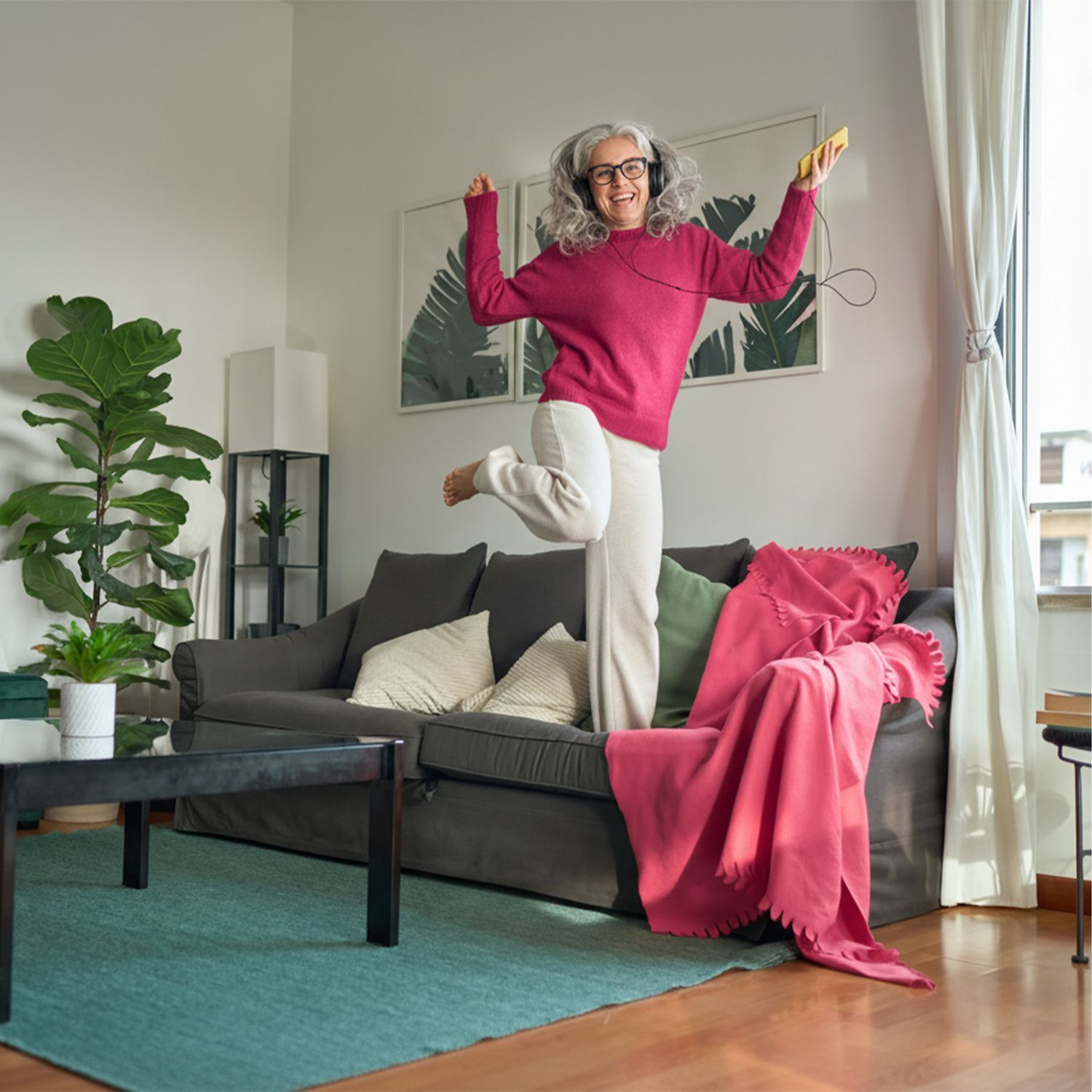 Der große Frühjahrsputz Person springt in einem hellen Wohnzimmer mit grauem Sofa, grünen und weißen Kissen, roter Decke und großem Fenster mit Blick auf Stadtgebäude.