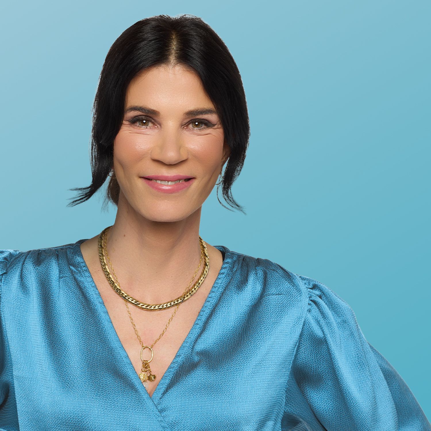 Frau mit schwarzem schulterlangem Haar trägt eine blaue Bluse mit Puffärmeln und zwei goldene Ketten um den Hals vor blauem Hintergrund