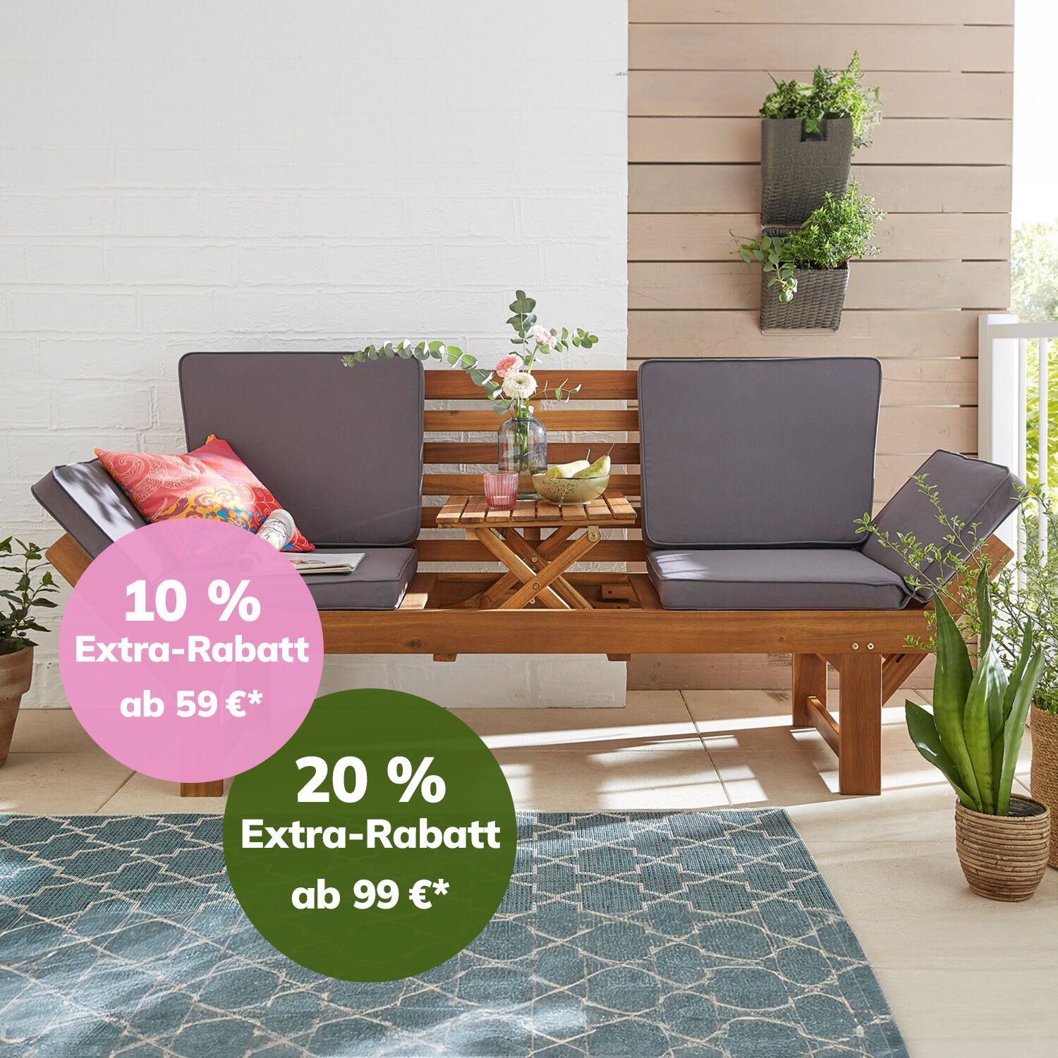 Zweisitzige Gartenbank mit grauen Polstern, bunten Kissen und kleinem Holztisch mit Dekoration auf hellem Boden vor weißer Wand und Pflanzen