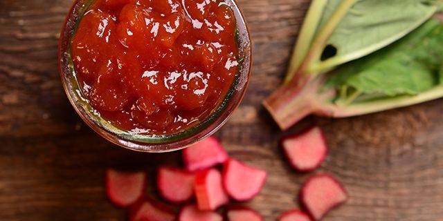 Rhabarber-Marmelade Glas mit rotem Rhabarberkompott auf Holztisch, daneben frische Rhabarberblätter und geschnittene Rhabarberstücke