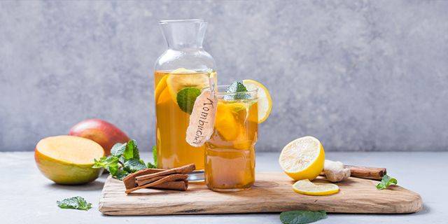 Kombucha Glaskaraffe und Glas mit bernsteinfarbenem Kombucha, garniert mit Zitronenscheiben, Minzblättern und Zimtstangen, vor grauem Hintergrund