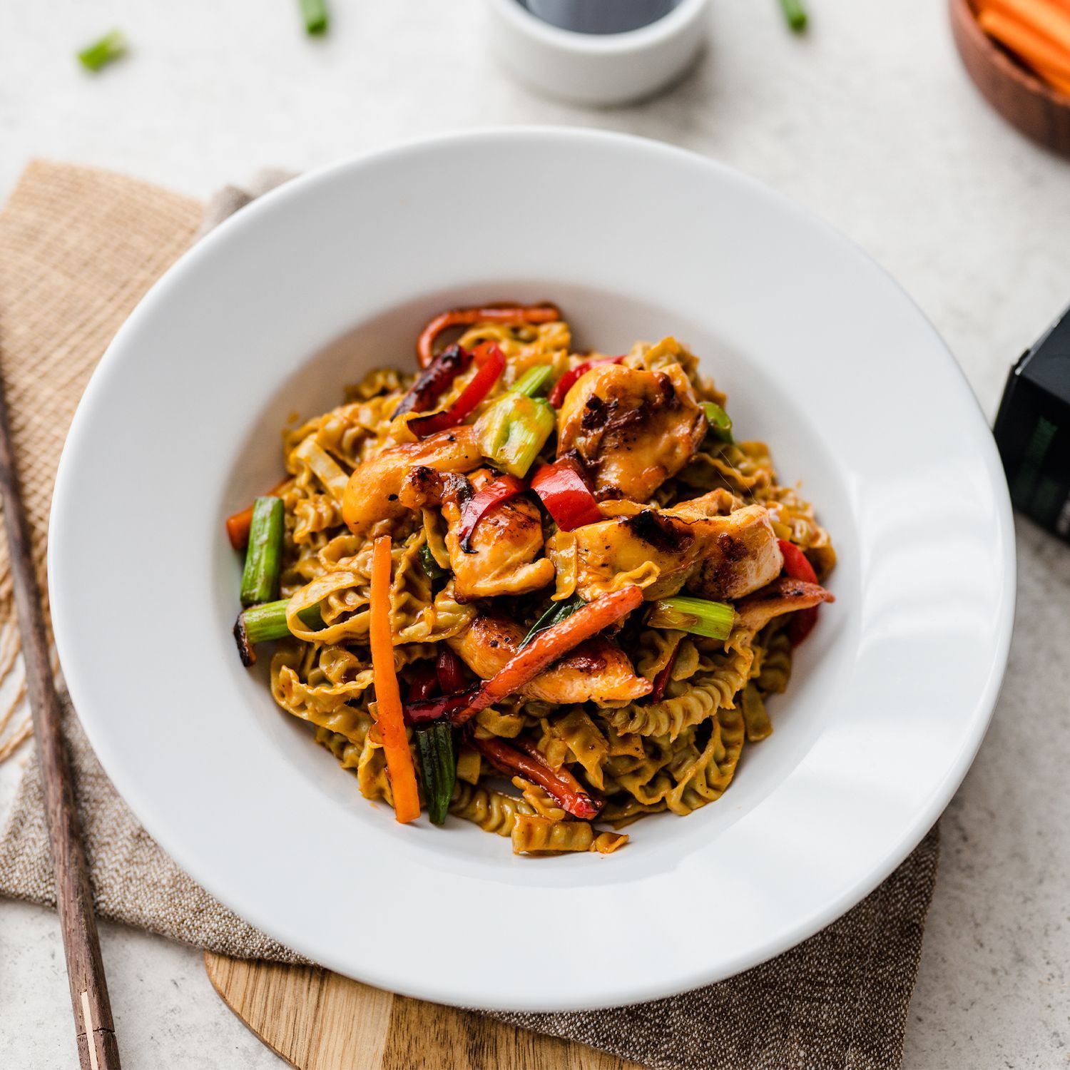 Bunte Gemüsepfanne mit Edamame-Nudeln, Erdnüssen und frischen Kräutern Teller mit gebratenen Nudeln, Karotten, Paprika und grünem Gemüse, daneben Just Taste Pasta, Essstäbchen und Sojasauce