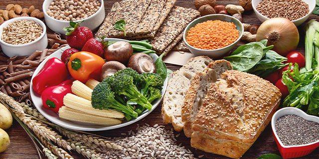 Ballaststoffreiche Lebensmittel für eine gesunde Darmflora Ein Tisch mit verschiedenen Lebensmitteln: Vollkornbrot, Gemüse wie Brokkoli, Paprika, Mais und Spinat, sowie Hülsenfrüchte und Nüsse in Schalen.