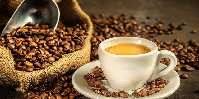 Kaffee Eine Tasse Kaffee auf einem Unterteller, umgeben von Kaffeebohnen. Im Hintergrund liegt ein Sack mit Kaffeebohnen und einem Schaufel.