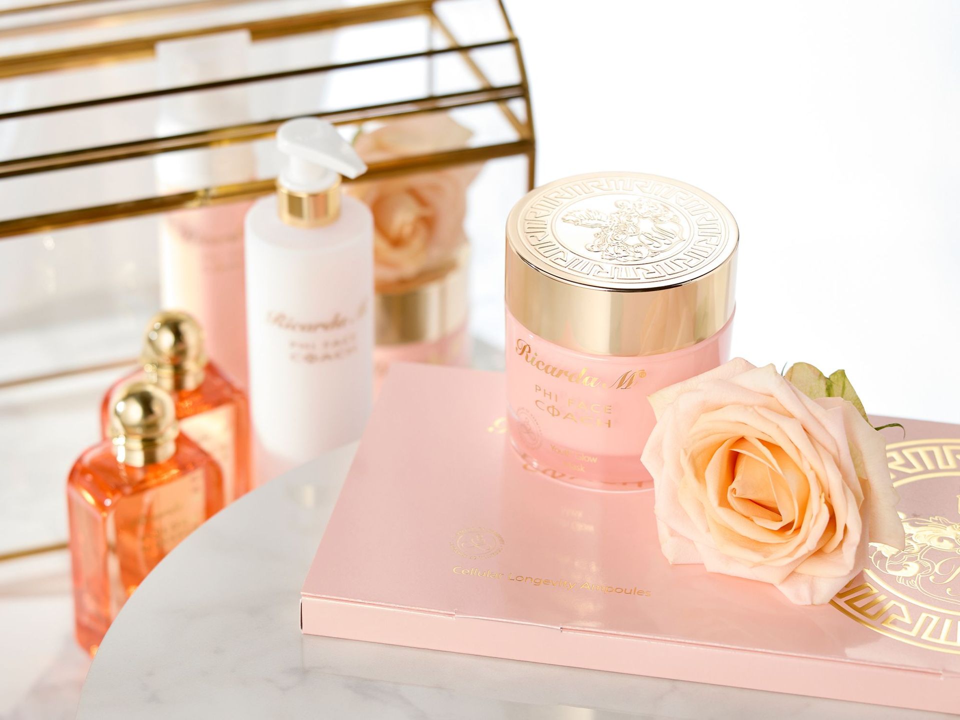 PHI FACE COACH – Entdecken Sie die innovative von Ricarda M! Pfirsichfarbene Kosmetikprodukte von Ricarda M., darunter ein Gesichtscreme-Tiegel mit goldfarbenem Deckel, eine Augencreme mit goldfarbenen Enden und eine Flasche, neben einer hellrosa Rose auf einem weißen Tisch.