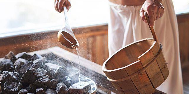 Sauna Aufguss Person gießt Wasser mit einer Kelle auf heiße Steine in einem Holzeimer, nur Oberkörper und Hände sichtbar.