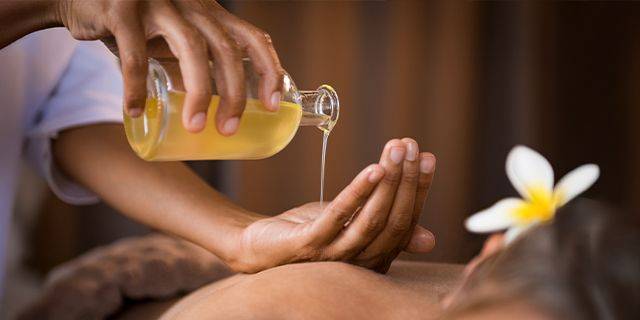 Sesamöl Hand gießt gelbliche Flüssigkeit aus Glasflasche in andere Hand, weiße Blüte im Hintergrund