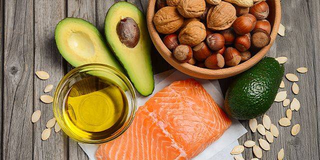 Omega-3 Halbierte Avocado, Schüssel mit verschiedenen Nüssen, Glas mit Olivenöl und rohes Lachsfilet auf Holzuntergrund.