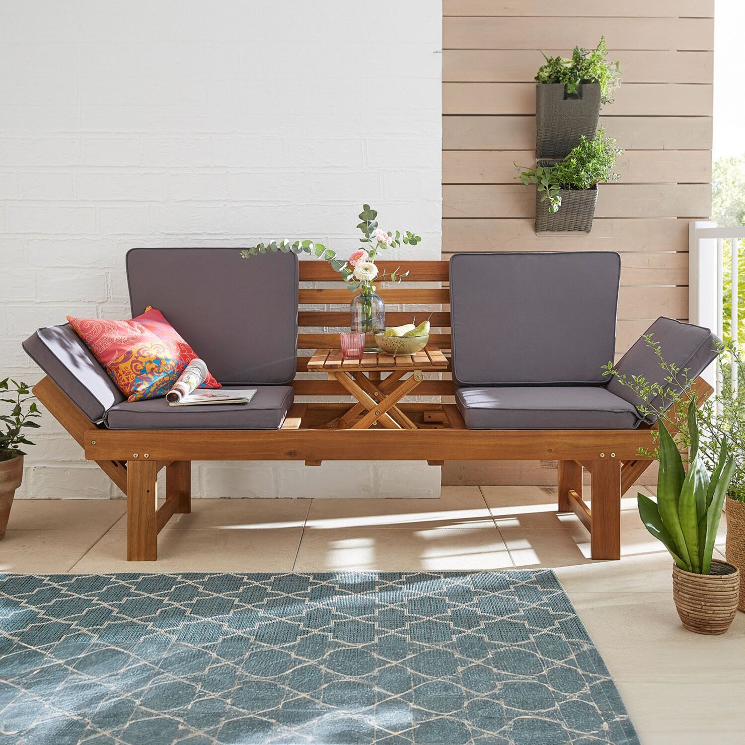 Gartenmöbel Holzbank mit grauen Polstern und bunten Kissen vor weißer Wand mit Pflanzen und kleinem Holztisch mit Vase