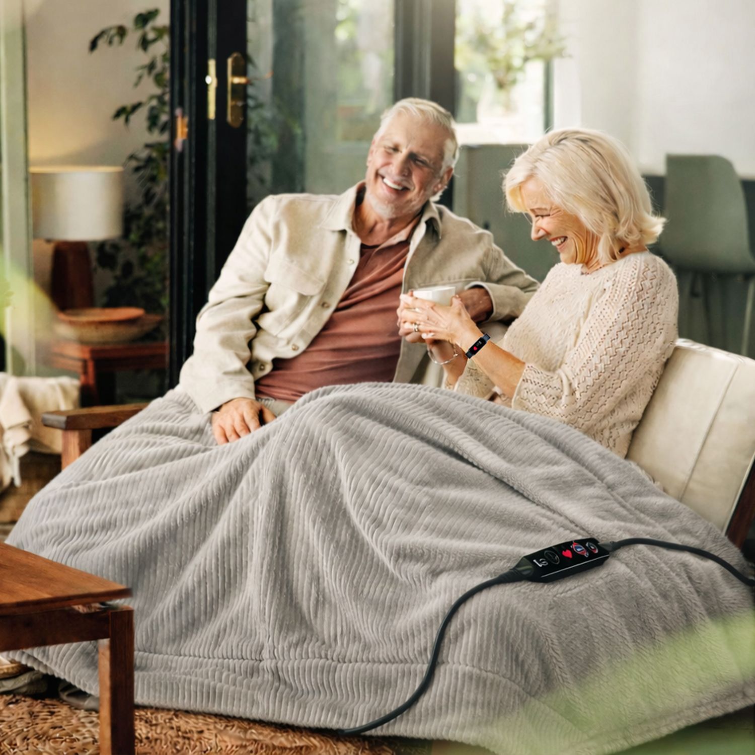 Beurer – Gesundheit und Wohlbefinden für Ihr Zuhause Älteres Paar sitzt auf Sofa unter einer grauen, gerippten Heizdecke mit Fernbedienung