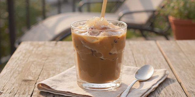 Vanille-Coffeeccino Glas mit Eiskaffee und Eiswürfeln auf einem hellen Stofftuch neben einem Löffel auf einem Holztisch im Freien