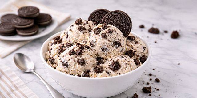 Vanille-Eiscreme mit Keksen Schale mit mehreren Kugeln Eis mit Keksstückchen und zwei ganzen Keksen daneben auf weißer Marmoroberfläche, daneben Löffel und Stapel Kekse