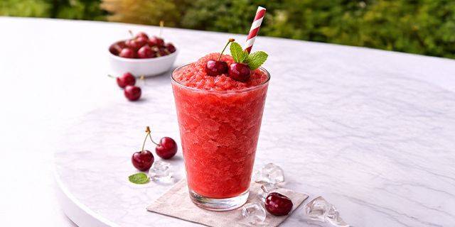 Kirsch-Slushie Glas mit rotem Slush-Eis, garniert mit Kirschen, Minzblatt und gestreiftem Trinkhalm auf weißer Marmoroberfläche mit Eiswürfeln und Kirschen im Hintergrund