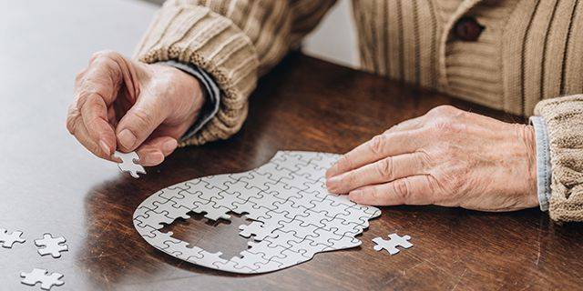 Demenz vorbeugen Hände einer älteren Person, die ein Puzzleteil über einem Puzzle in Form eines menschlichen Kopfes halten