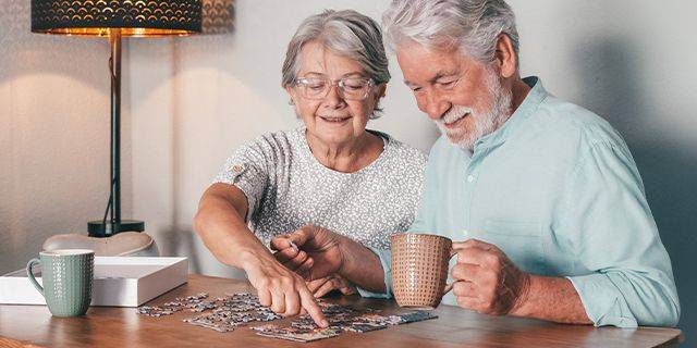 Demenz vorbeugen Älteres Paar sitzt am Tisch und legt gemeinsam ein Puzzle, die Frau zeigt auf ein Puzzleteil, der Mann hält eine Tasse in der Hand