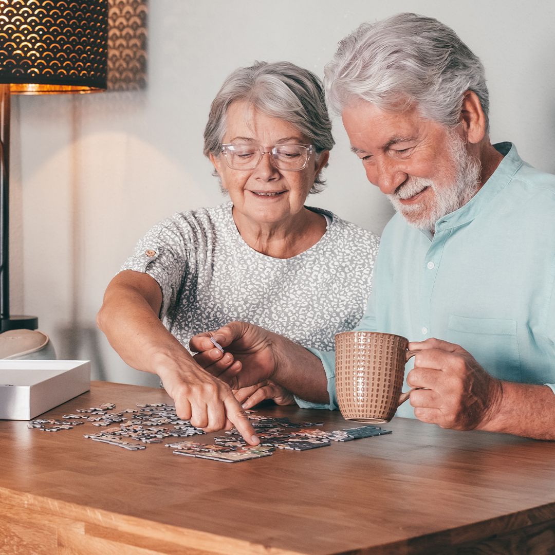 Demenz Älteres Paar sitzt am Tisch und legt gemeinsam ein Puzzle, die Frau zeigt auf ein Puzzleteil, der Mann hält eine Tasse in der Hand