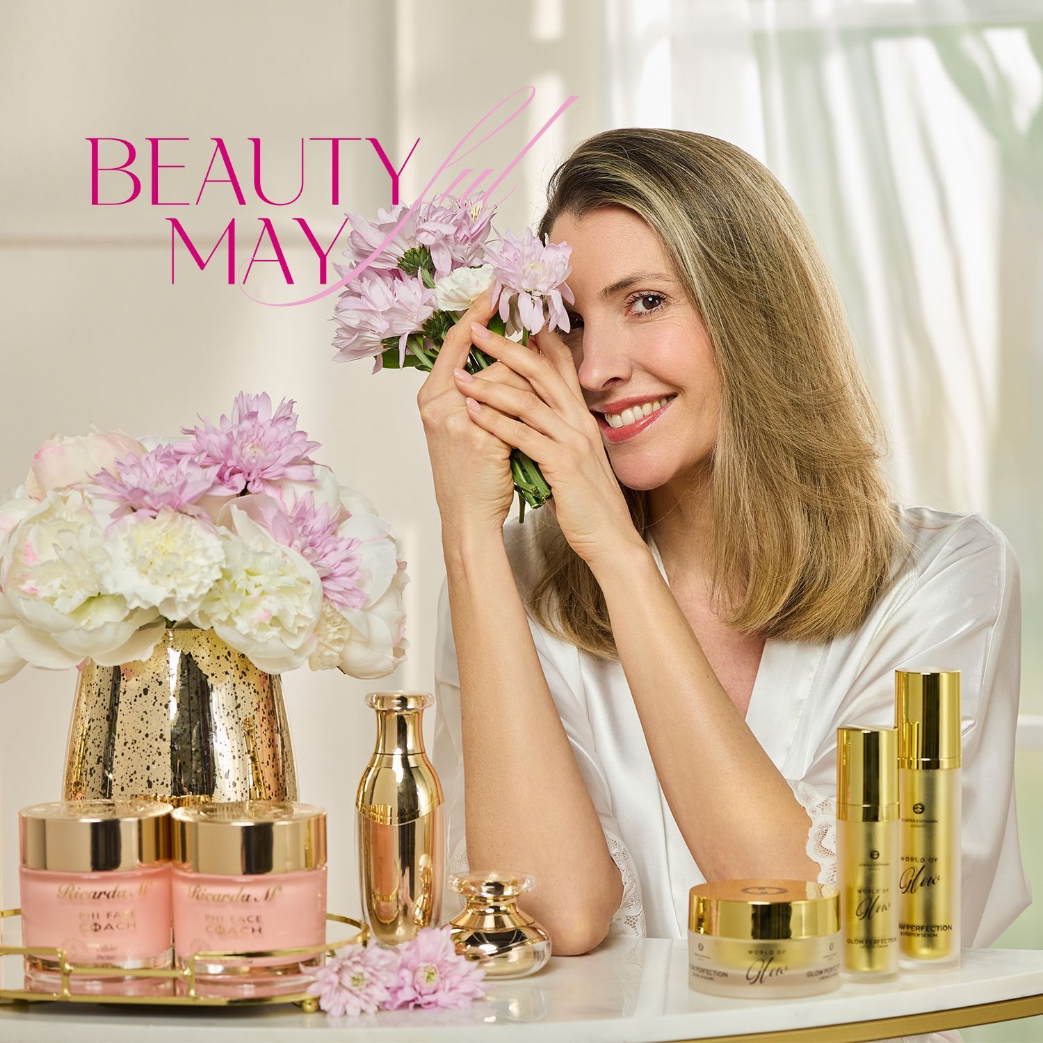 Beautyful May Frau mit mittellangen blonden Haaren in weißem Satinoberteil hält rosa Blüten vor Gesicht, davor auf Tisch goldene und rosa Kosmetikbehälter sowie weiße und rosa Blumen in goldener Vase