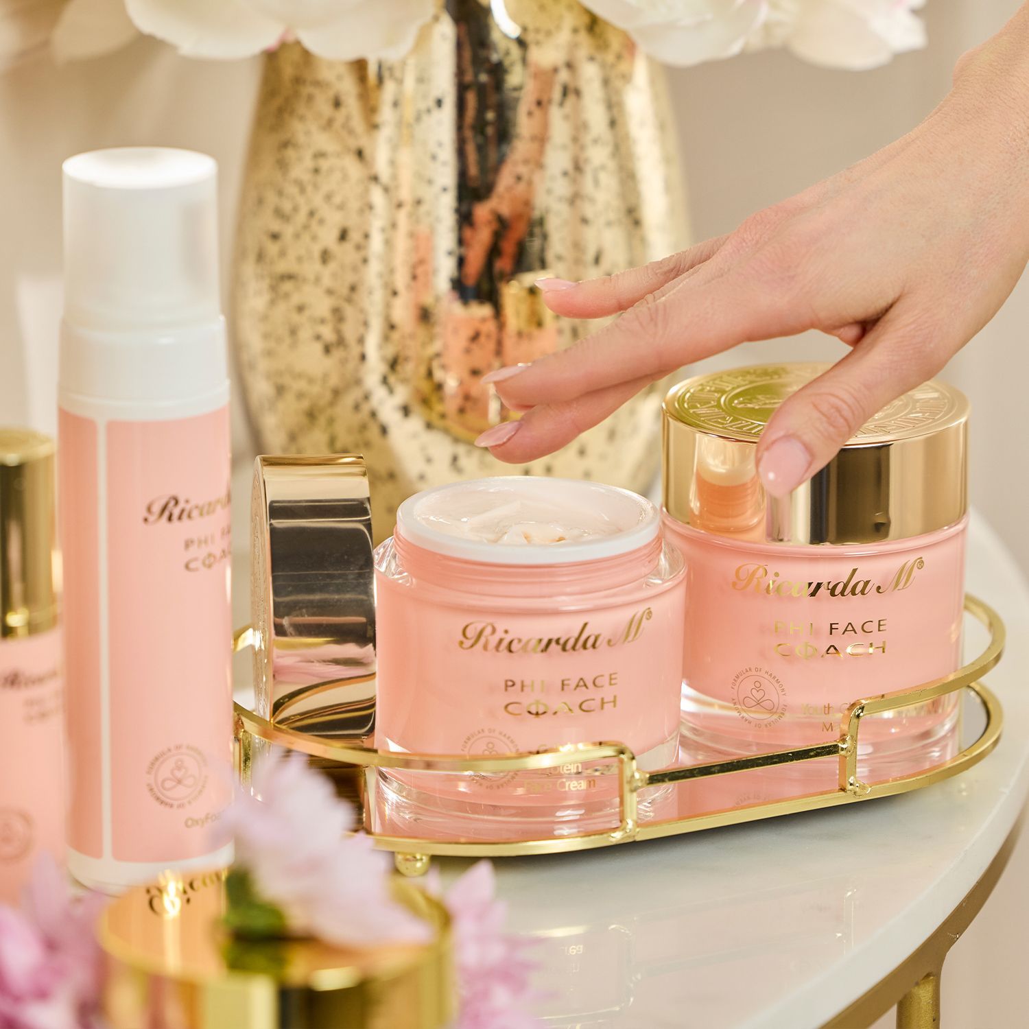 PHI FACE COACH – Entdecken Sie die innovative von Ricarda M! Hand greift nach einem geöffneten, rosafarbenen Gesichtscremetiegel mit goldfarbenem Deckel auf einem goldfarbenen Tablett neben weiteren Kosmetikprodukten
