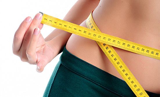Ausschnitt des Bauchs einer Frau, sie hält sich ein gelbes Maßband um ihren Bauch um ihren Bauchumfang zu messen.