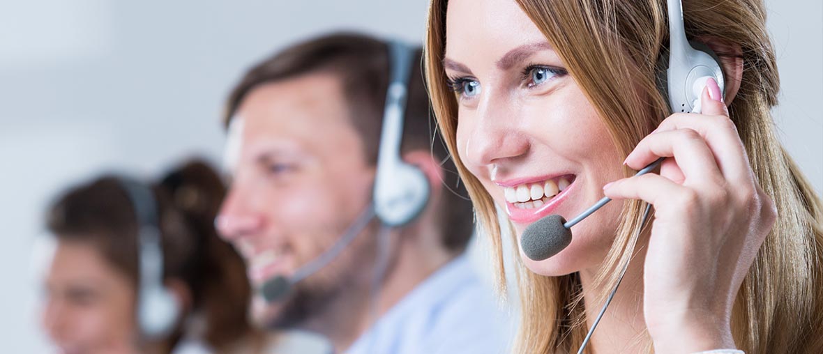 Junge Frau im Callcenter mit Headset. Im Hintergrund zwei Herren, ebenfalls im Callcenter tätig, leicht verschwommen.